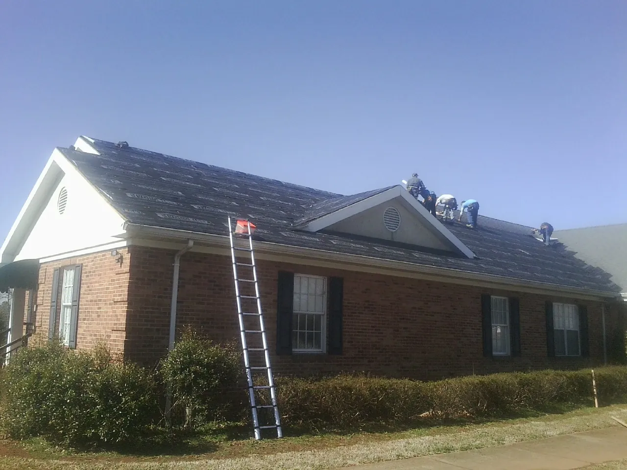Crew tearing shingles off a brick ranch roof with a ladder set — Metal Roof Repair work by our crew in Barton