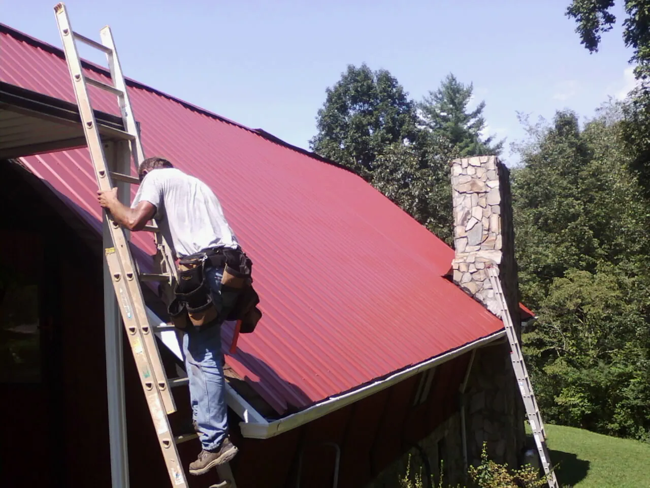 Roofer on a ladder beside a red metal roof and stone chimney — Soffit & Fascia Repair by Roofing Brothers in Barton